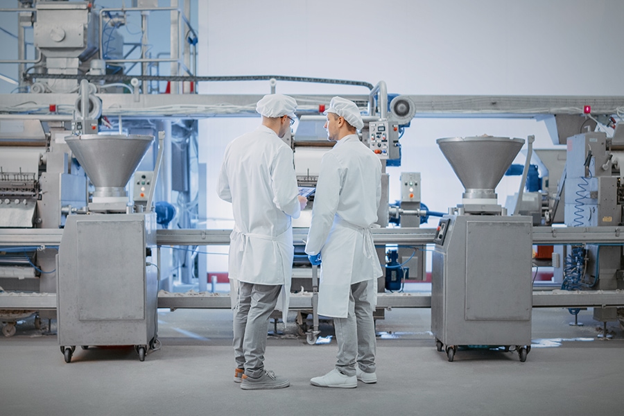 Two Young Food Factory Employees Discuss Work-Related Matters. Male Technician or Quality Manager Uses a Tablet Computer for Work. They Wear White Sanitary Hat and Work Robes.