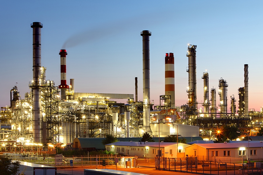 An industrial refinery complex is brightly lit in the evening, with numerous tall chimneys and structures visible against the twilight sky