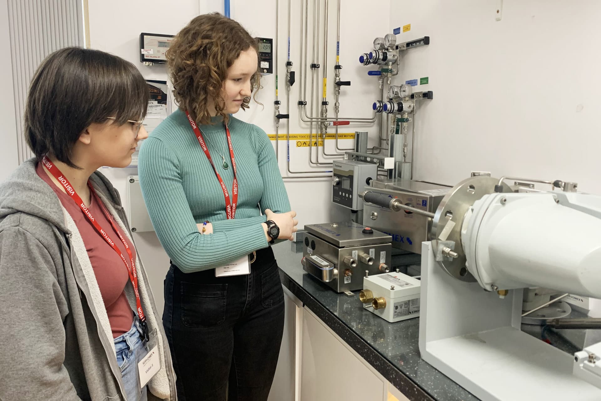 Two women apprentices in a laboratory examining a complex machine with focused expressions and collaborative spirit.