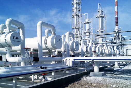 Industrial facility with large white pipelines, pressure vessels, and tall metal towers arranged in rows under a blue sky, showing complex processing equipment typical of a petrochemical or gas plant.