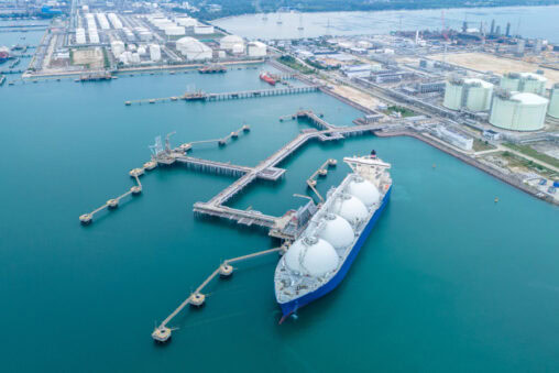 An aerial view of a large LNG tanker docked at a loading terminal, surrounded by long piers and industrial storage tanks along the shoreline.